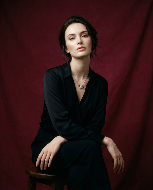 Woman in a black dress sitting on a wooden stool against a red curtain and wearing heart necklace from Swarav Creations.